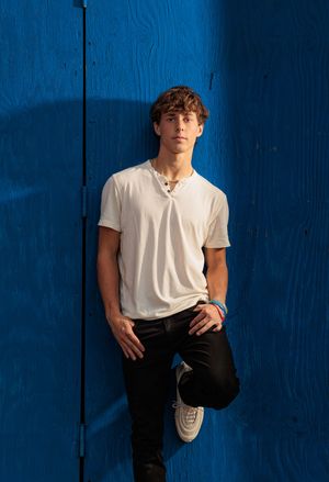 Young man in white t-shirt and black jeans leaning against vibrant blue wooden wall.