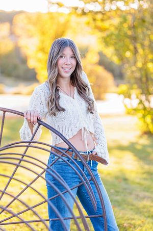 Young woman in cream crochet top and jeans poses with rustic metal wheel in golden autumn outdoor setting. Gilmanton NH.