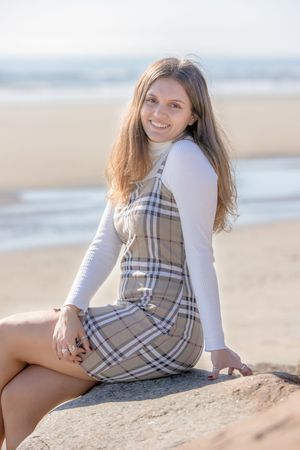 Smiling teenage girl in plaid dress sitting on rock at sunny beach with ocean backdrop. Wells Reserve at Laudholm, Wells ME.