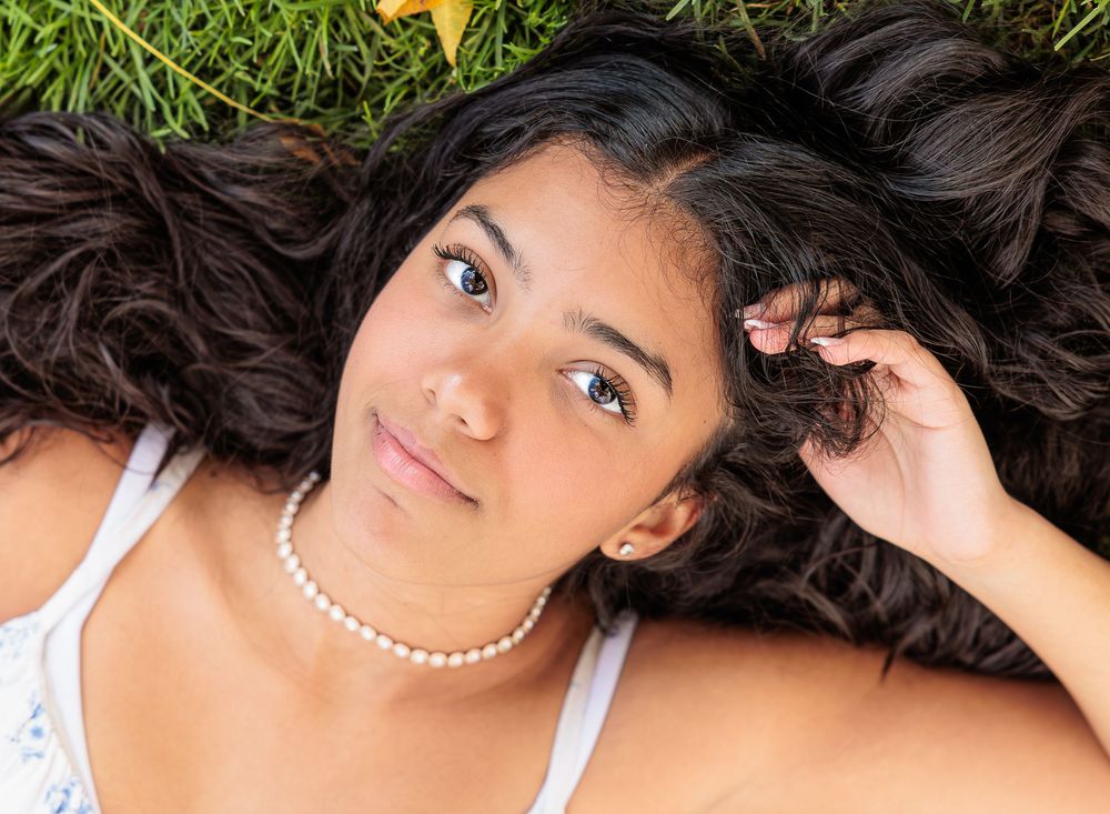 Young woman with long dark hair and pearl necklace lies on green grass looking up at camera. Cate Park Wolfeboro NH