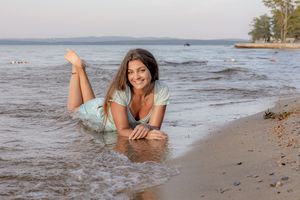 Smiling young woman in light blue dress lying on sandy shoreline at lake's edge. Ellacoya State Park, Gilford NH.