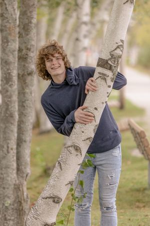 Smiling teenage boy with curly hair hugging a birch tree in a tree-lined park. Cotton Valley Rail Trail, Wolfeboro NH