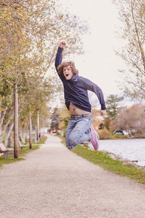 Energetic teenage boy jumping in air with fist raised on scenic tree-lined pathway. Cotton Valley Rail Trail, Wolfeboro NH