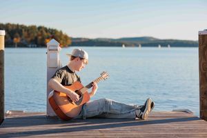 Teenage boy playing acoustic guitar while sitting on wooden dock beside a calm lake. Cate Park Wolfeboro NH