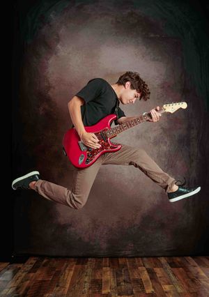 Teen boy jumping while playing red electric guitar on dark backdrop.