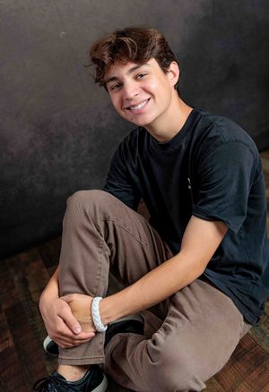 Smiling teen boy in black shirt sitting casually on studio floor.