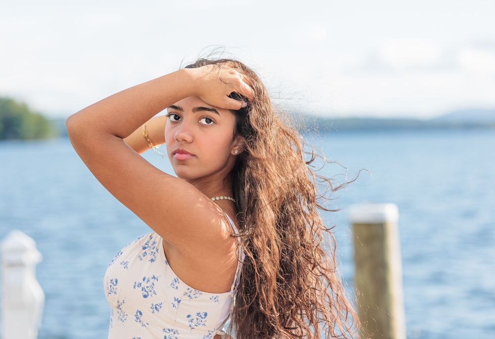 Young woman with curly hair shields eyes from sun while posing near sparkling blue lake outdoors. Cate Park, Wolfeboro NH.