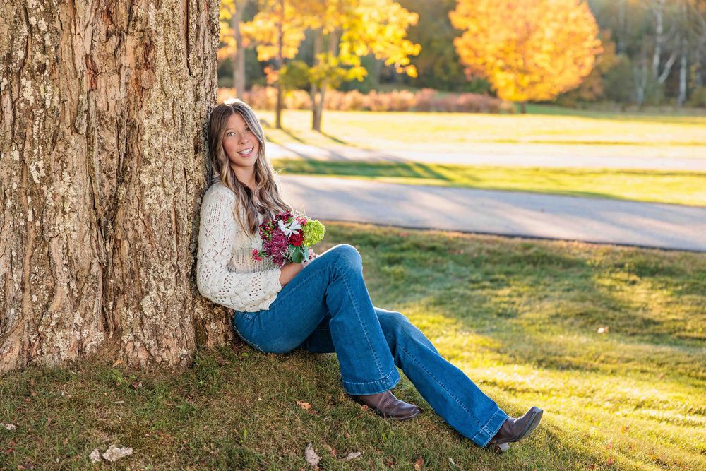 Smiling young woman in cream sweater and jeans sits against large tree holding flowers in fall park. Gilmanton NH.