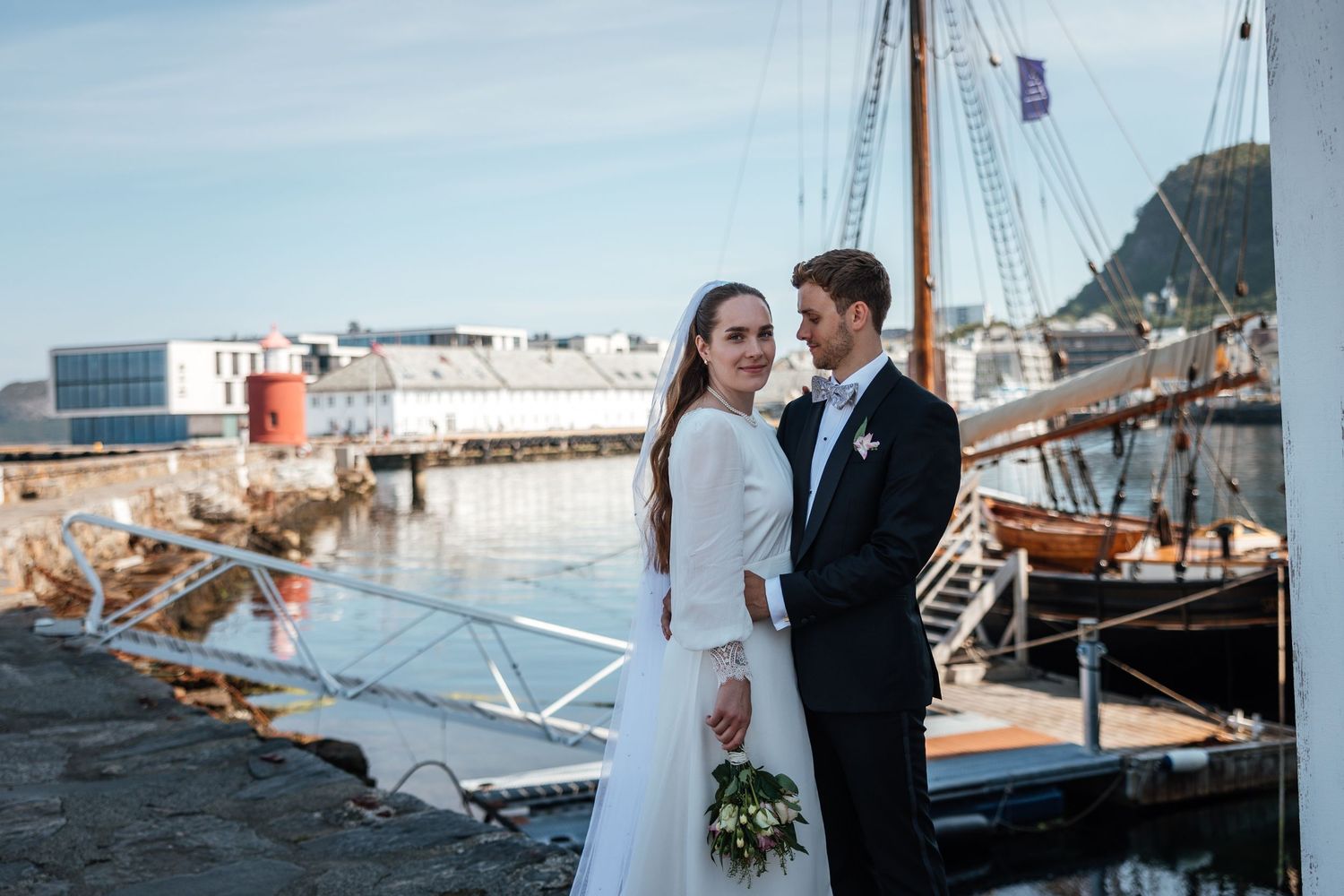 Bride and groom share a romantic moment on a waterfront pier with a scenic Norwegian coastal town and hillside in the background.