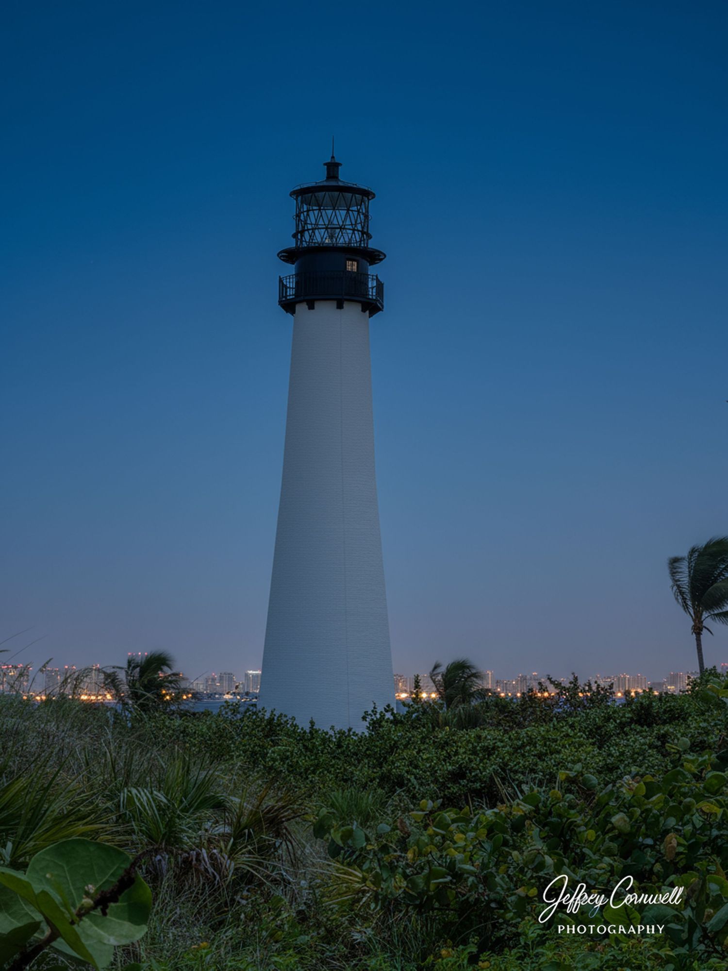 White lighthouse silhouetted against deep blue sky with city lights glowing in the background.
