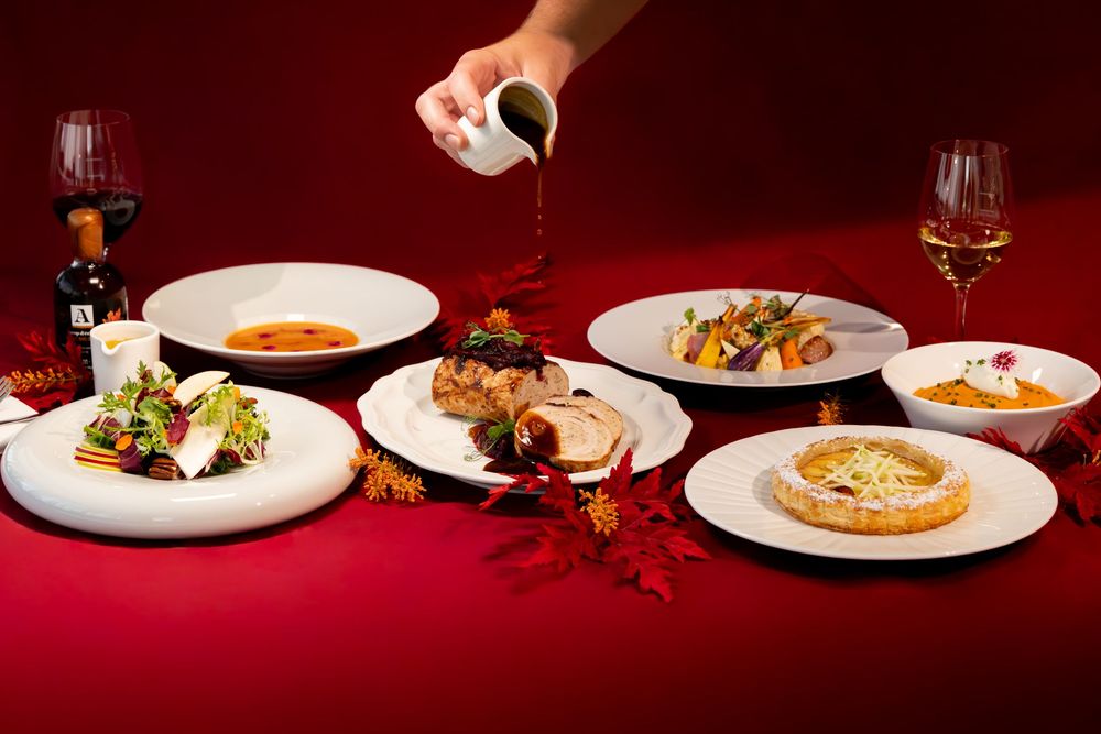 Elegant dinner spread with multiple plates of gourmet dishes on a red tablecloth with wine glasses and sauce being poured.