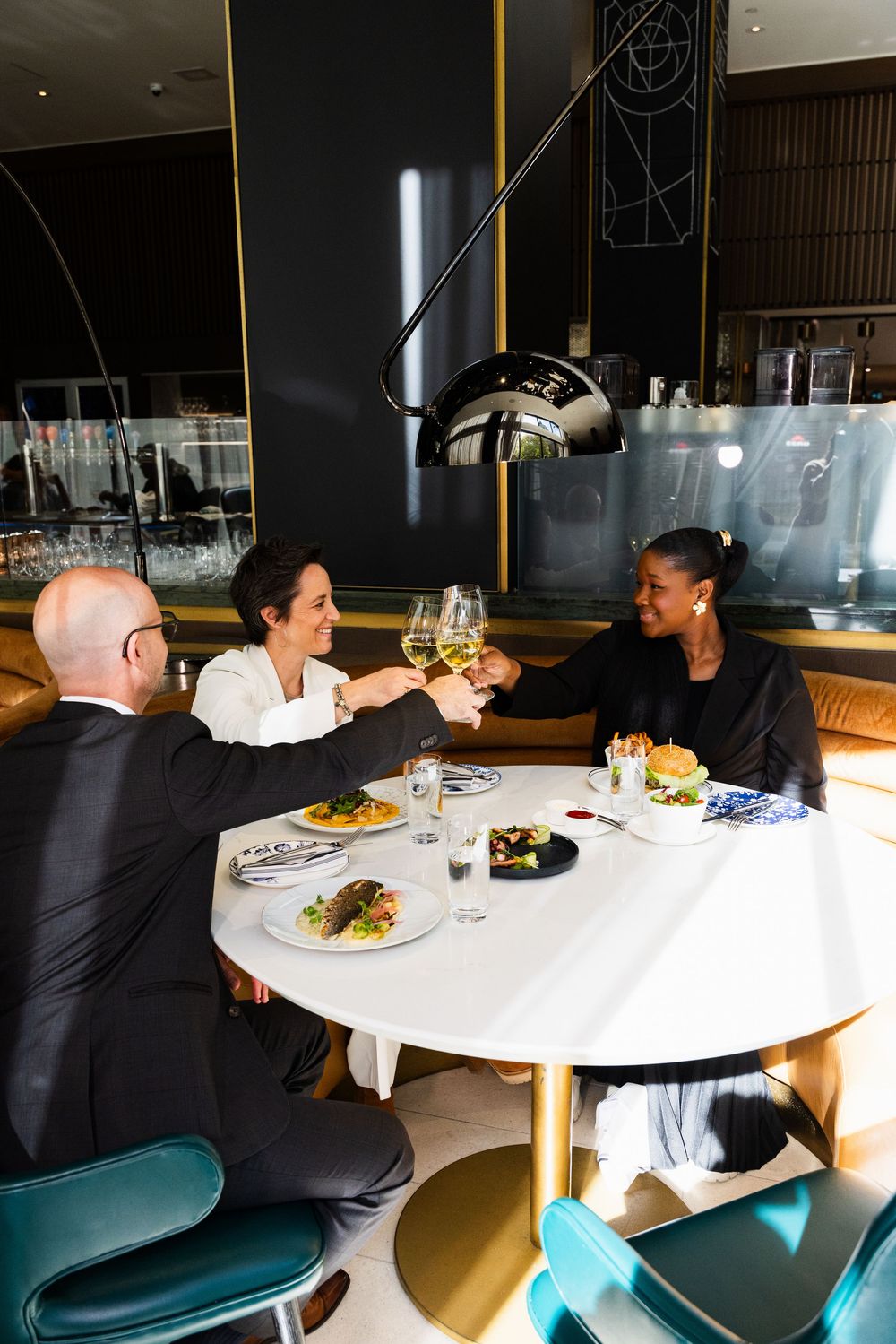 Diners seated at a modern white round table in an upscale restaurant with teal velvet chairs.