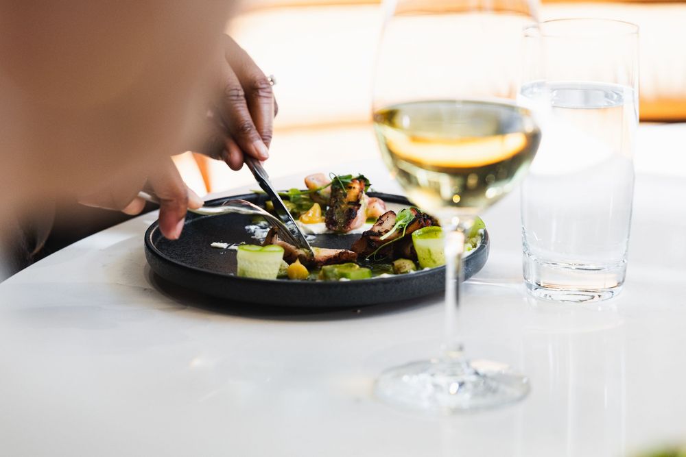 Hands serving a fresh salad on a black plate with a glass of white wine on a white tablecloth.
