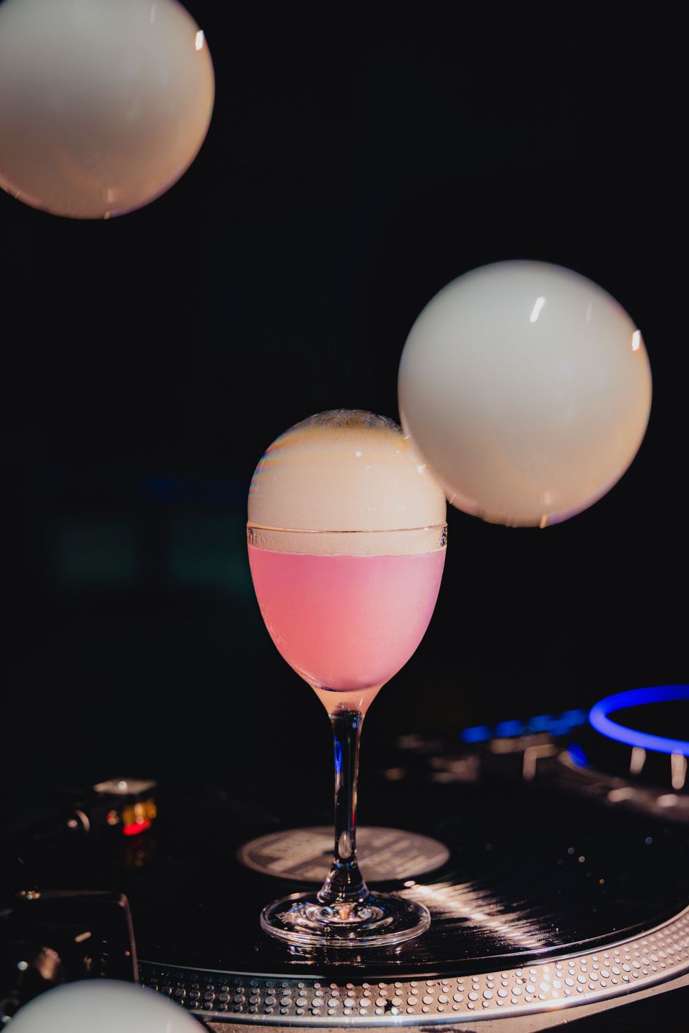 A pink cocktail with white foam sits on a turntable with white balloons floating above in a dark setting.