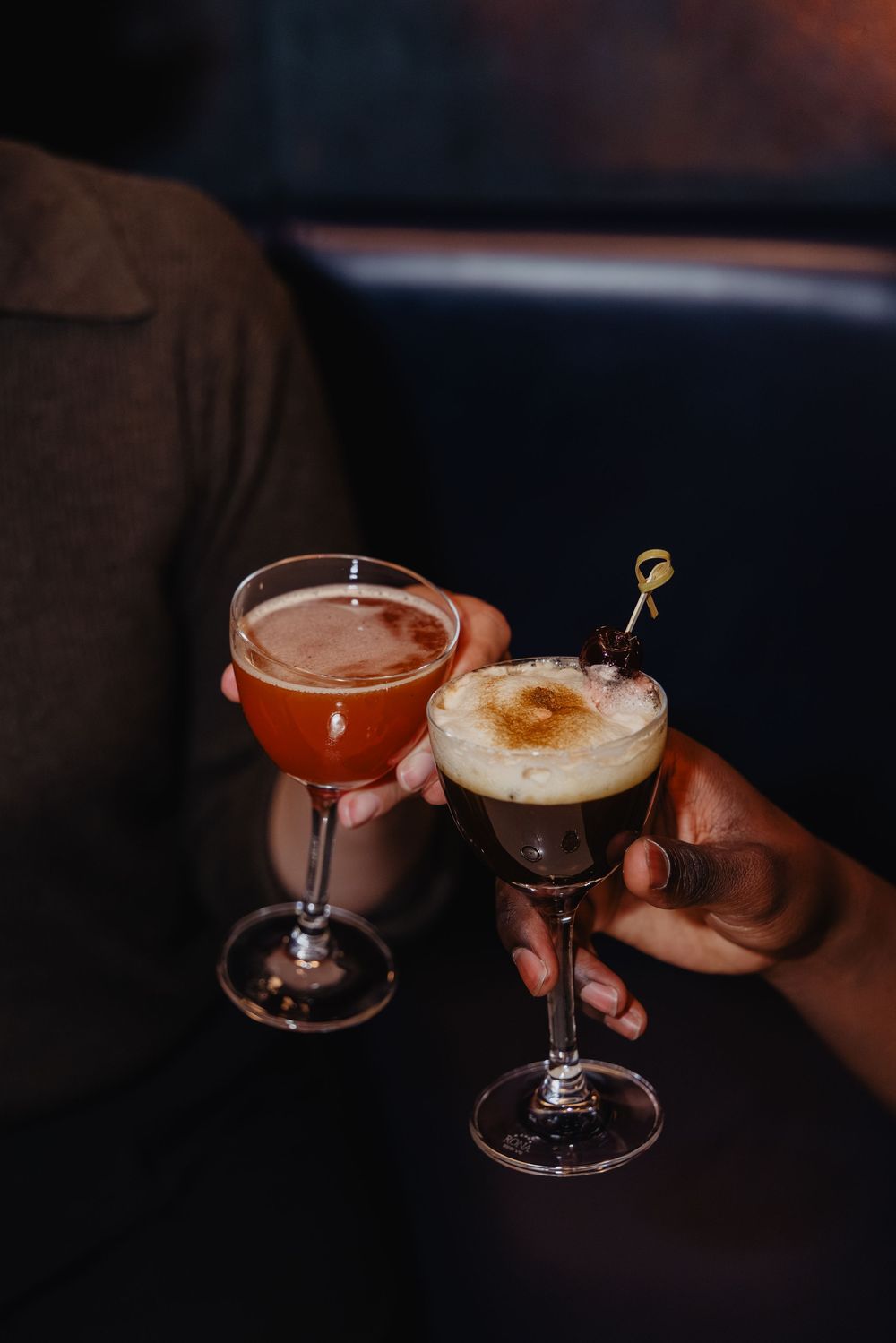 A hand holds an elegant cocktail glass with a frothy espresso martini against a dark moody background.