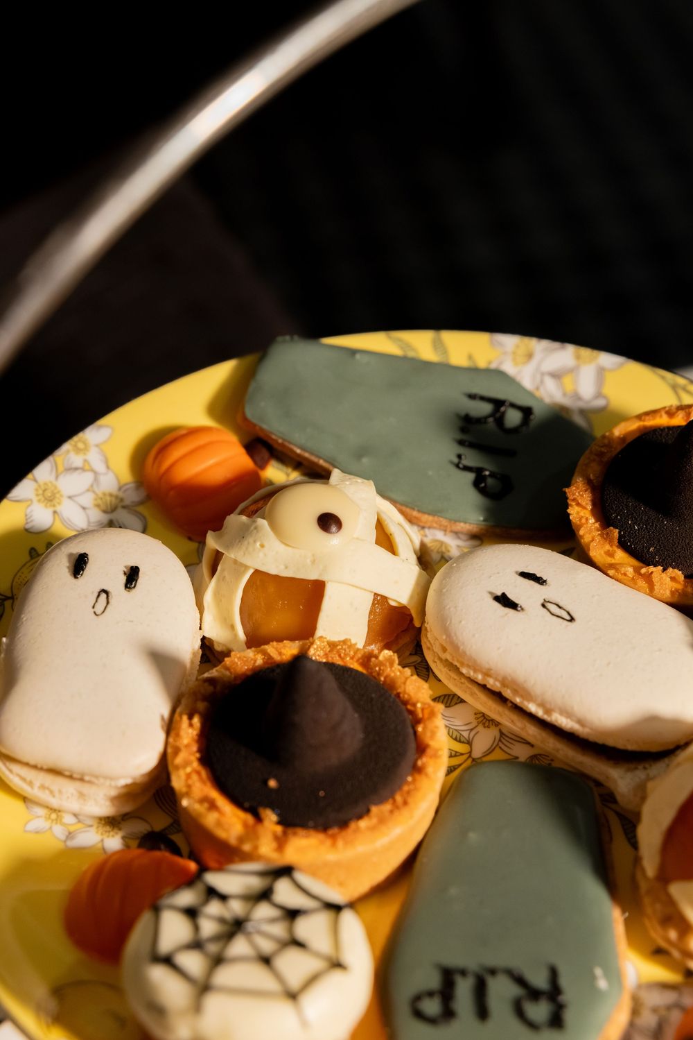 Halloween themed desserts including ghost macarons, chocolate tarts, and spooky decorated cookies on a yellow plate.