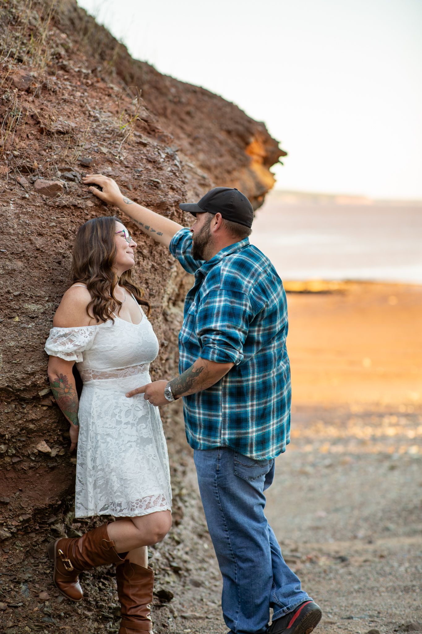 Couple poses together against rocky cliff at sunset with woman in white dress and man in plaid shirt.