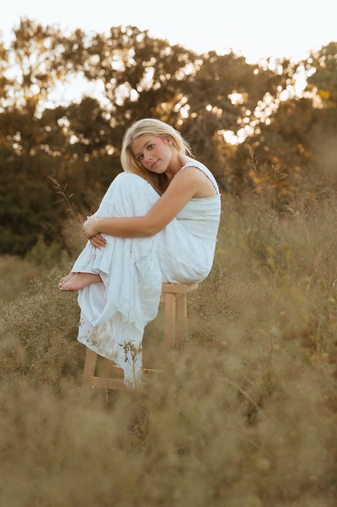 A dreamy golden hour portrait of someone in a flowing white dress sitting on a wooden stool in a wild grass field.