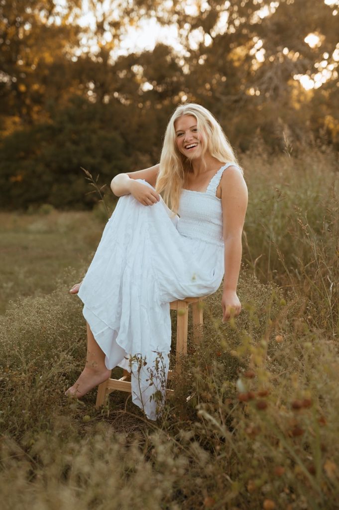 A joyful outdoor portrait of someone in a white sundress sitting on a wooden stool amidst tall grass at sunset.