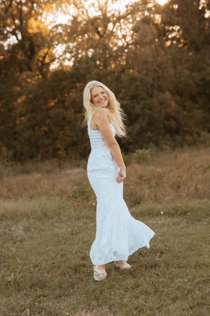 A flowing white sundress twirls gracefully in a golden sunset field with trees in the background.