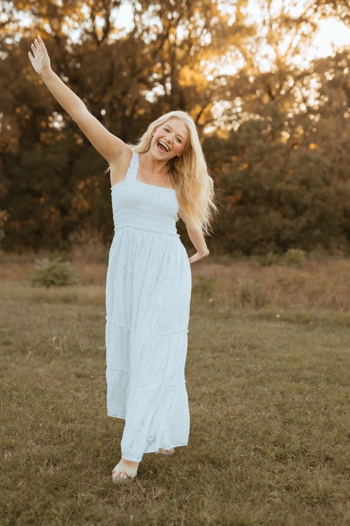 A joyful person in a white summer dress twirls and dances in a grassy field at golden hour.