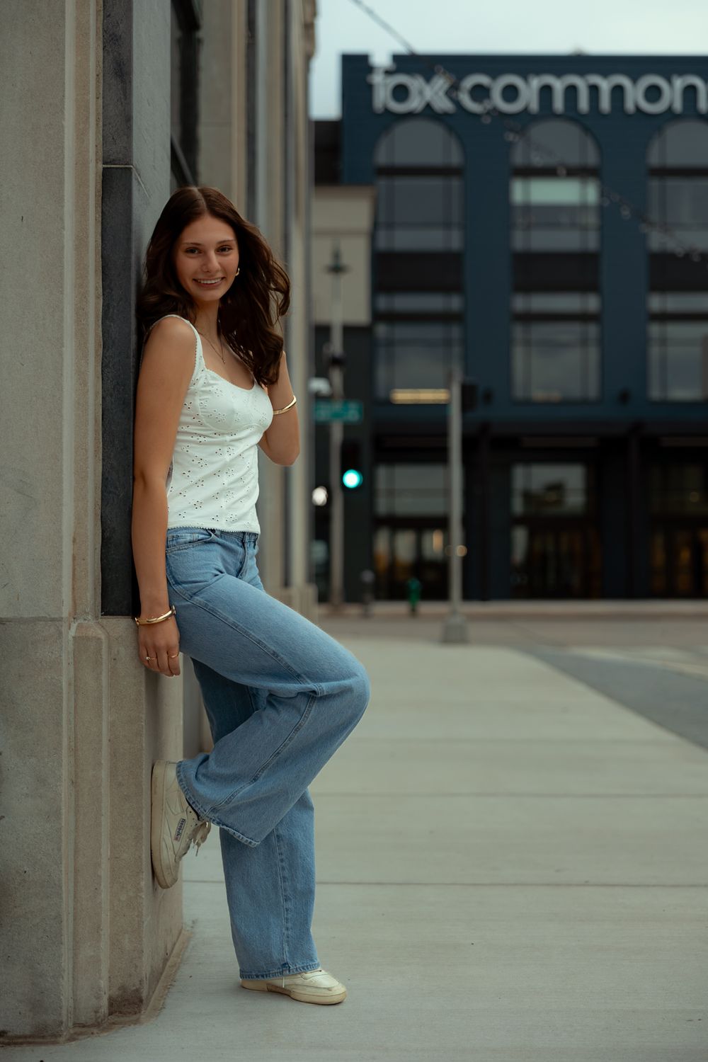 A person in a white tank top and baggy jeans poses playfully against a concrete column outdoors.
