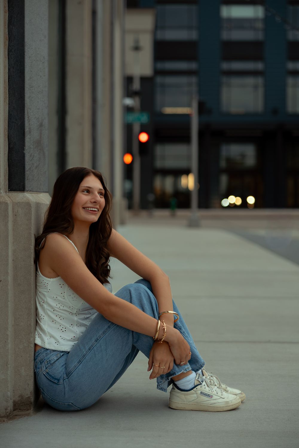 Person in jeans and white sneakers sits casually on concrete steps in an urban setting at dusk.
