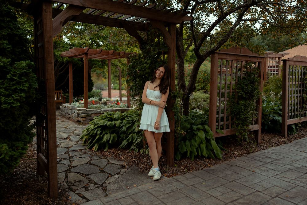 A person in a light dress stands by a wooden trellis and stone wall in a garden setting.