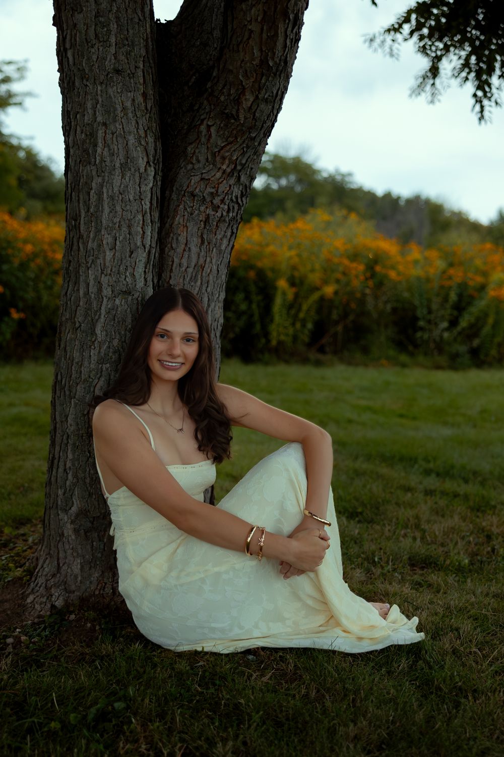 A person in a white dress sits gracefully against a tree trunk in an outdoor setting with autumn foliage in the background.