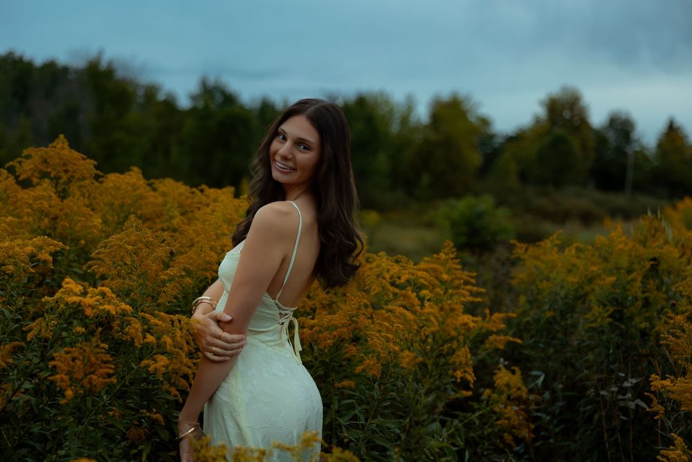 A woman in a white dress poses among vibrant yellow wildflowers in a scenic outdoor setting during golden hour.