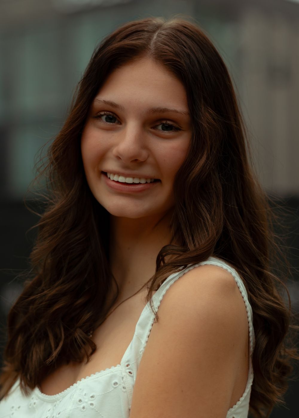 A portrait of someone with long brown hair wearing a white sleeveless top, smiling warmly at the camera.