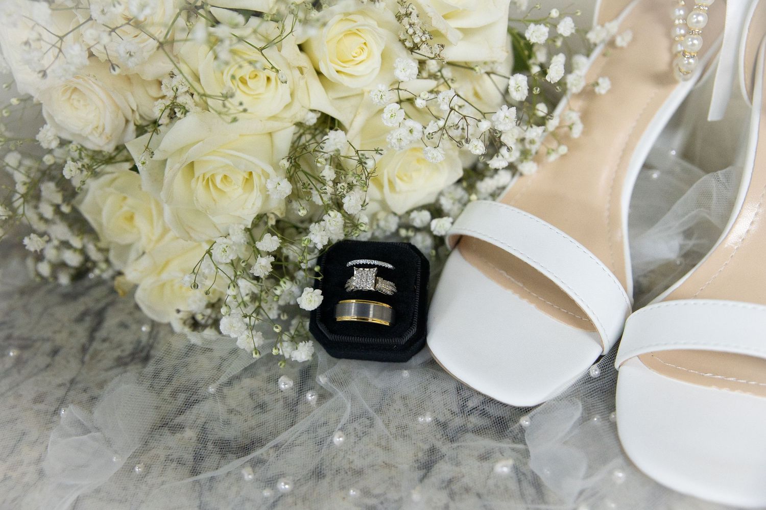 A delicate white rose and baby's breath wedding bouquet rests beside elegant bridal shoes and accessories.