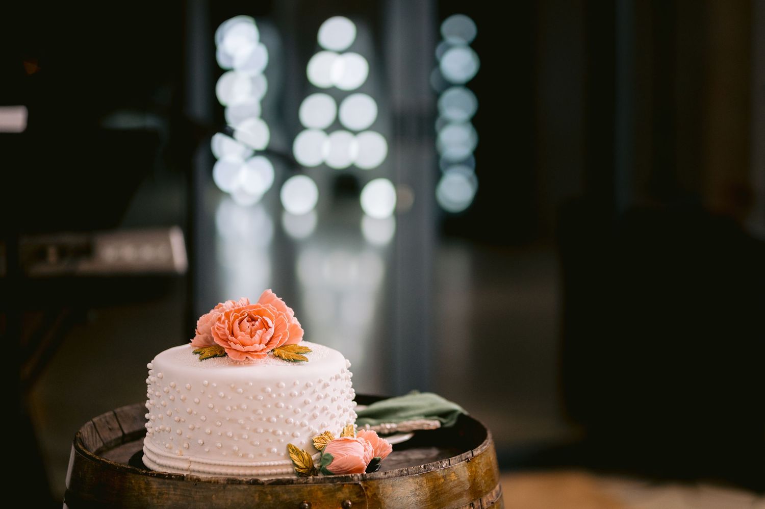 Elegant white wedding cake with peach rose decoration sits on rustic wooden table in dimly lit venue.