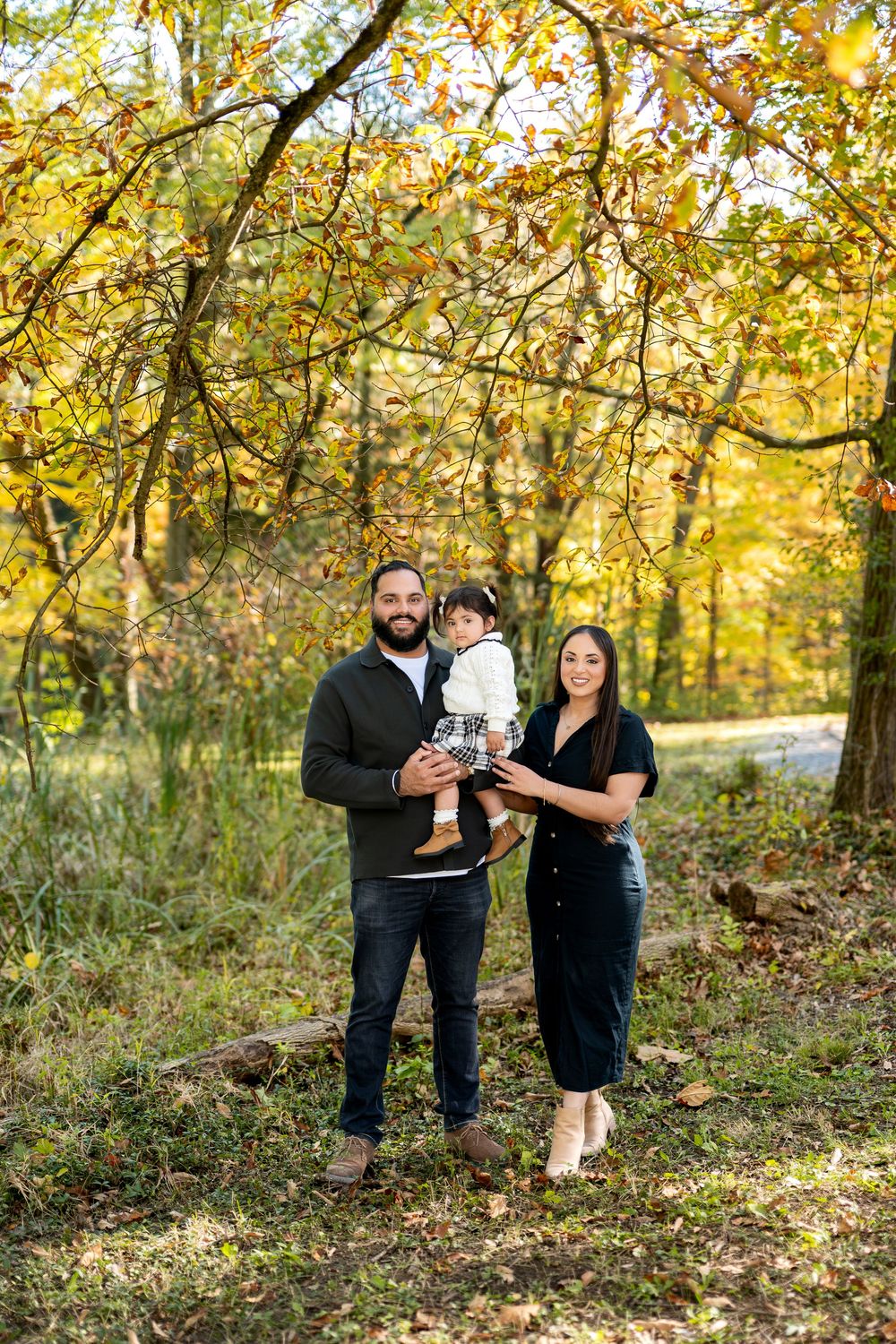 Family of three posing together under golden fall foliage in a wooded park.