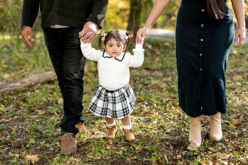 Toddler girl in plaid skirt holds parents hands while walking outdoors in autumn park.