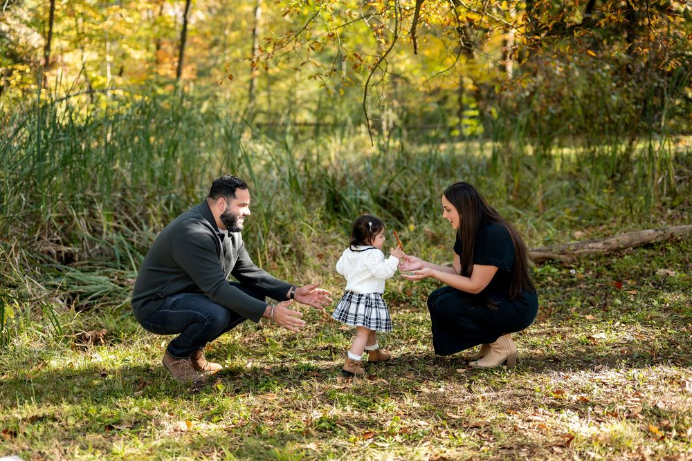 Young family playing with toddler girl in a sunlit forest with fall foliage.