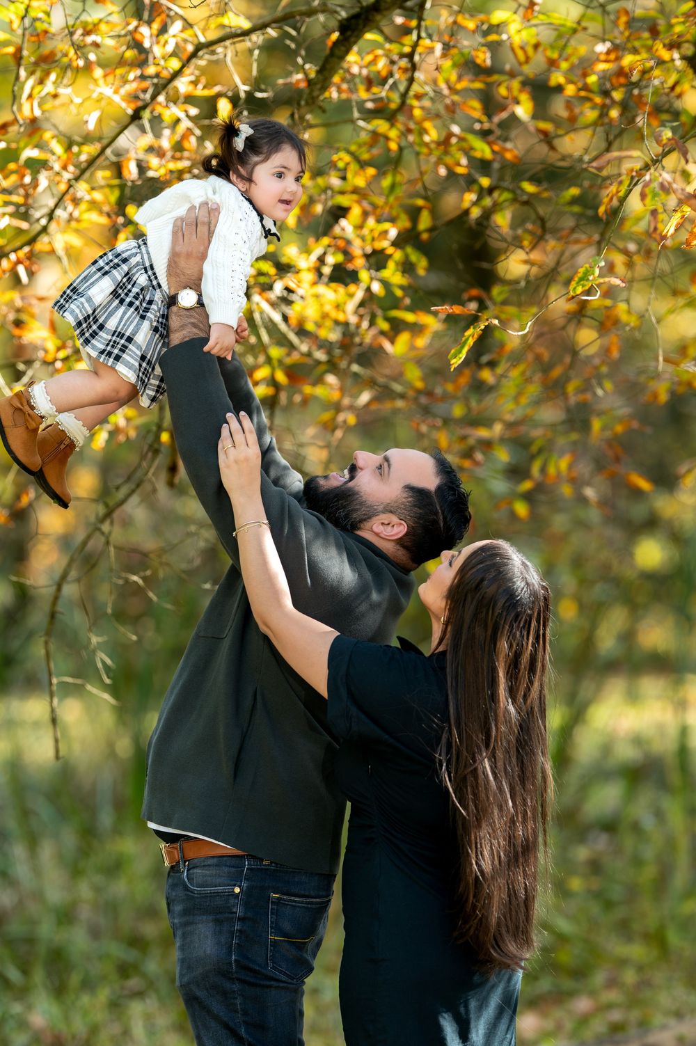 Father lifting smiling toddler girl in the air while mother watches in autumn park.