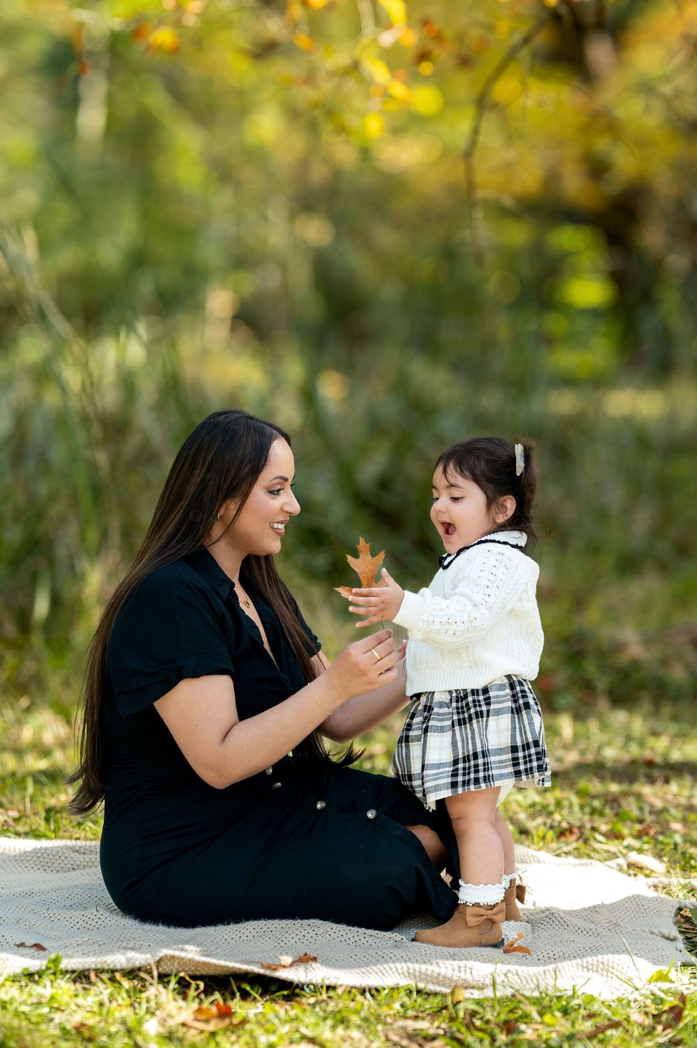 Mother sitting on blanket showing autumn leaf to curious toddler daughter in park.
