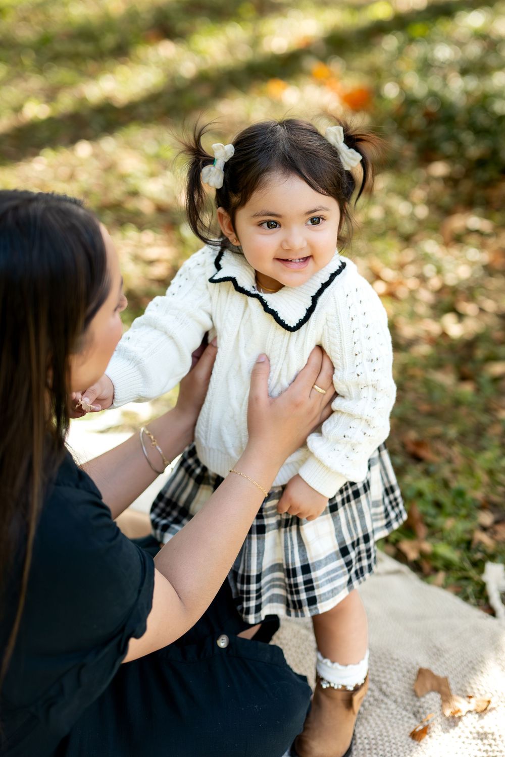 Adorable smiling toddler girl in white sweater held by mother outdoors in fall park.