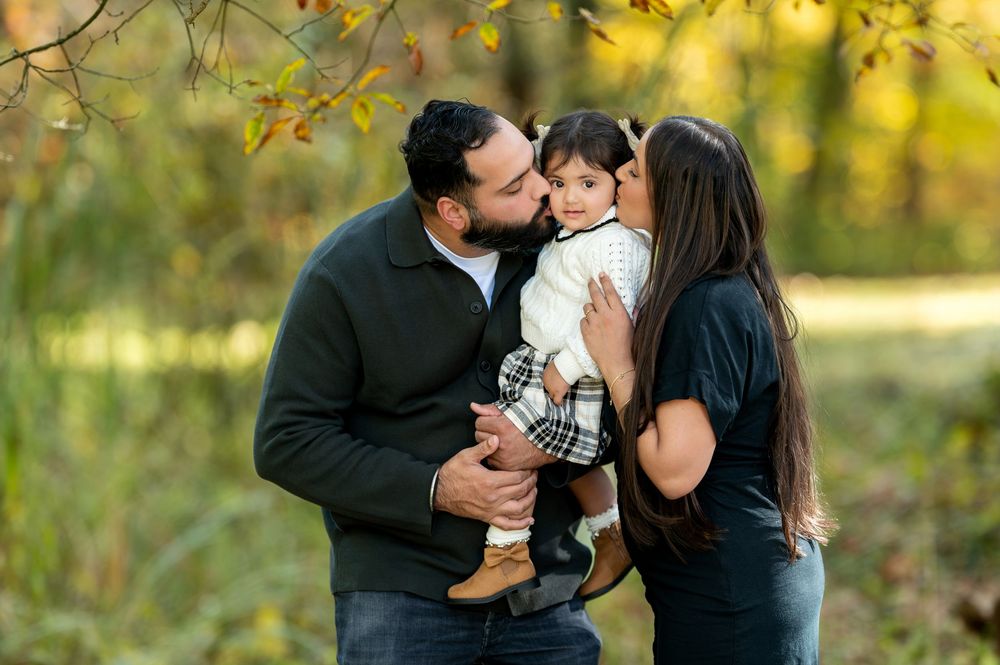Parents sharing a kiss on their toddler daughters cheeks in fall foliage park.