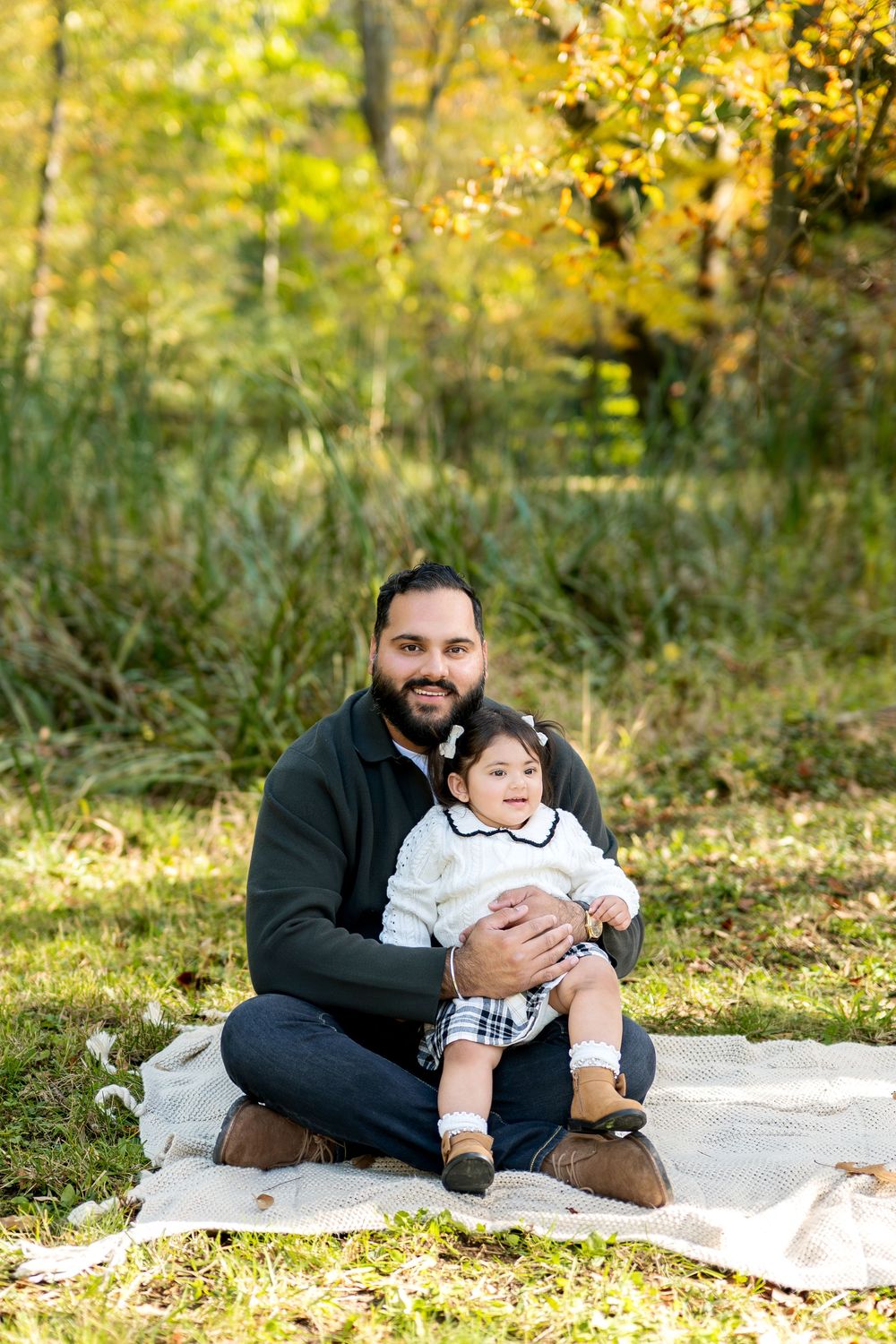 Father sitting cross-legged on blanket in autumn park holding baby in white sweater and plaid skirt.