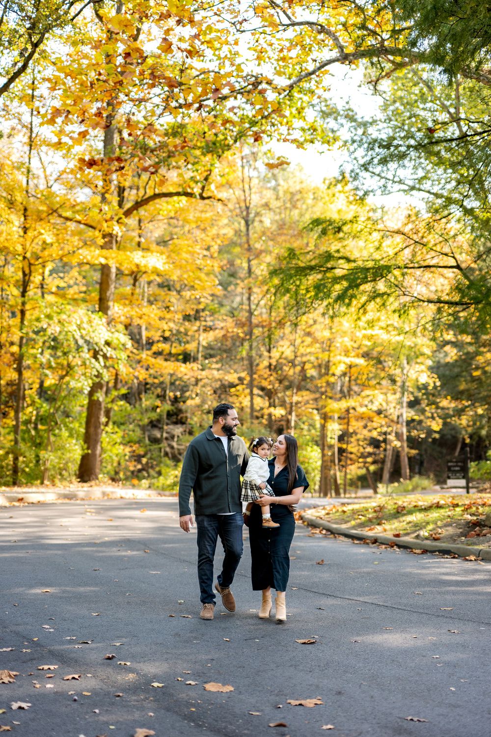 Happy family of three walking on road amid golden autumn trees, mother holding toddler.