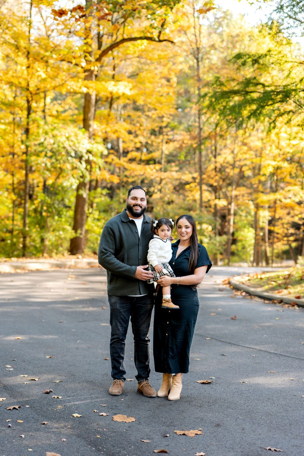 Family of three posing on road with stunning yellow fall foliage in background.