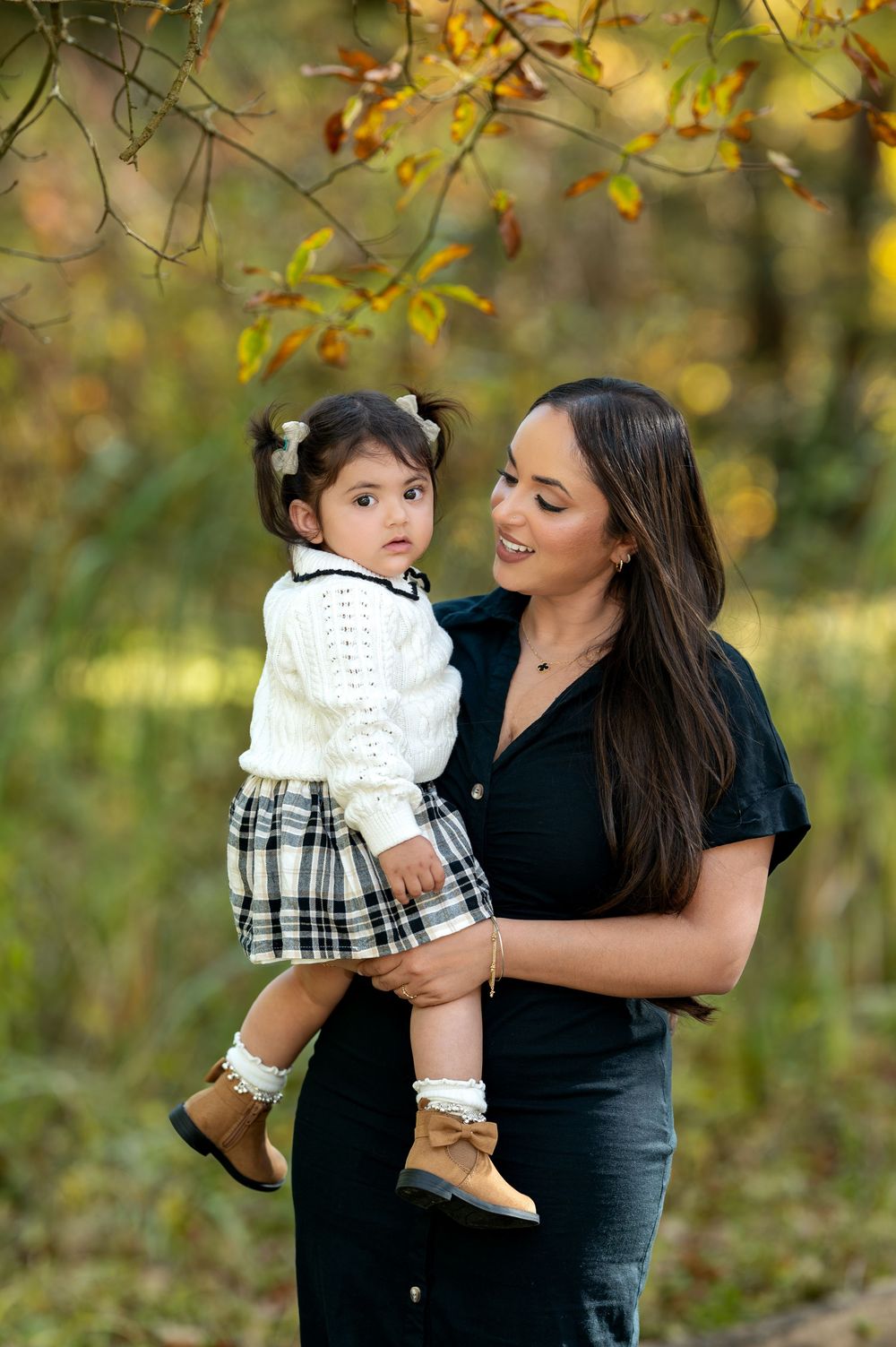 Smiling mother holding toddler girl in white sweater and plaid skirt in autumn park.