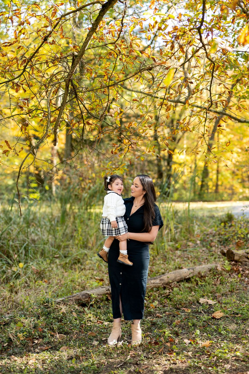 Happy mother lifting laughing toddler daughter under colorful autumn tree branches.