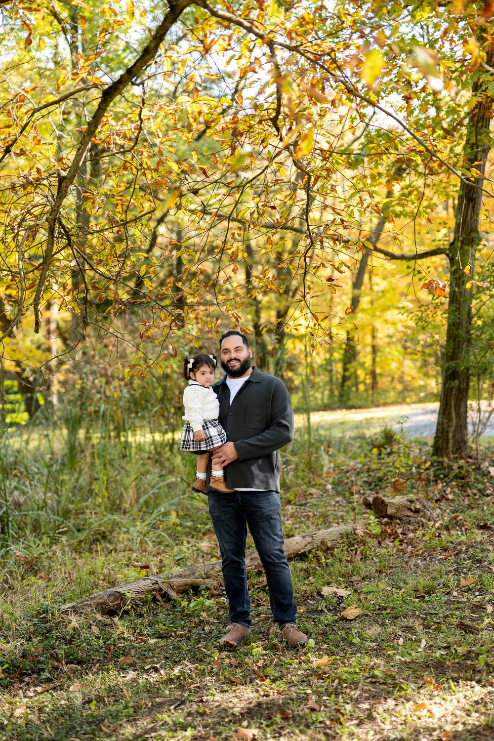 Bearded father holding toddler daughter in autumn woodland setting with golden leaves.