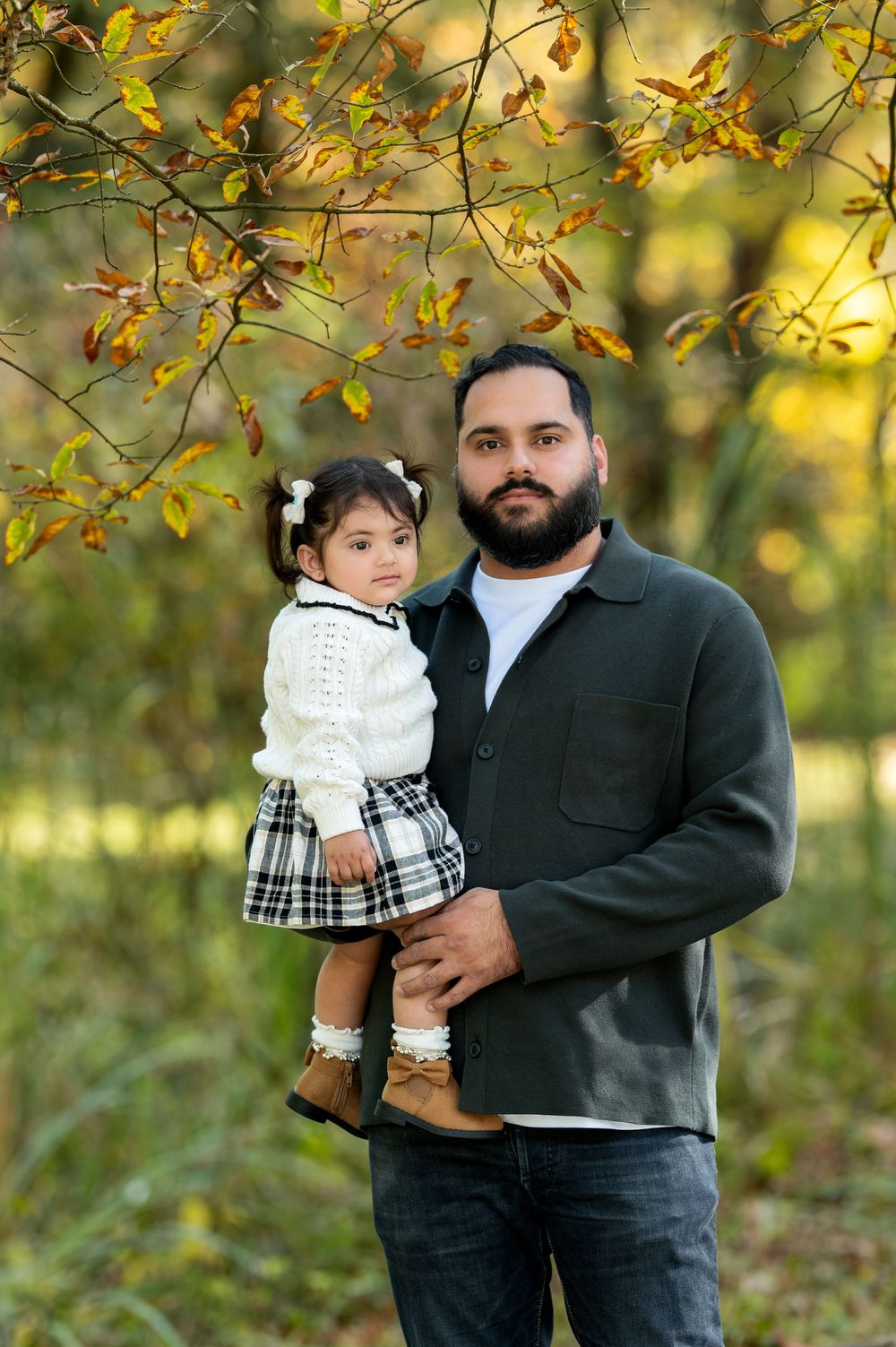 Father with beard holding young daughter in plaid skirt under fall foliage.