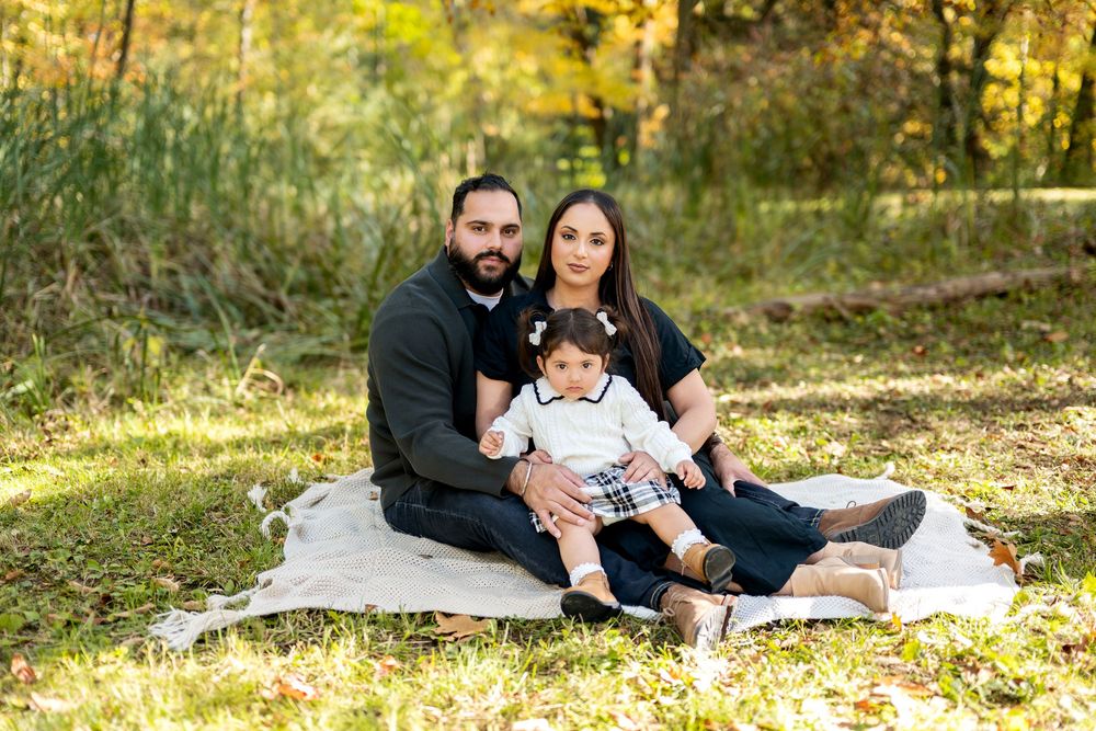Family of three sitting on blanket together in sunny autumn park setting.