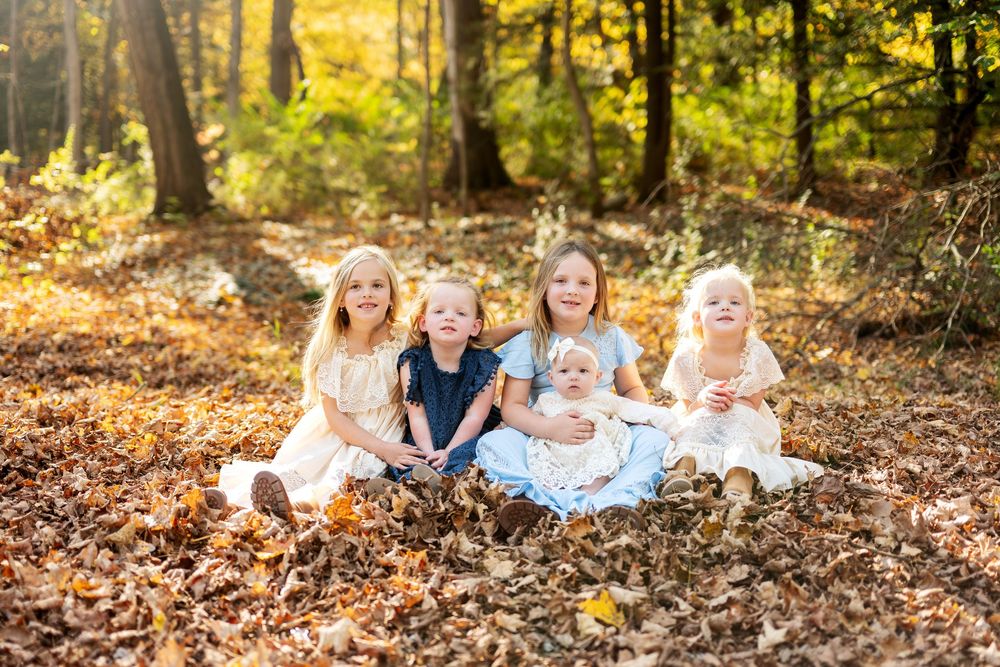 Five young siblings sit together smiling in a pile of fallen autumn leaves in a sunlit forest.