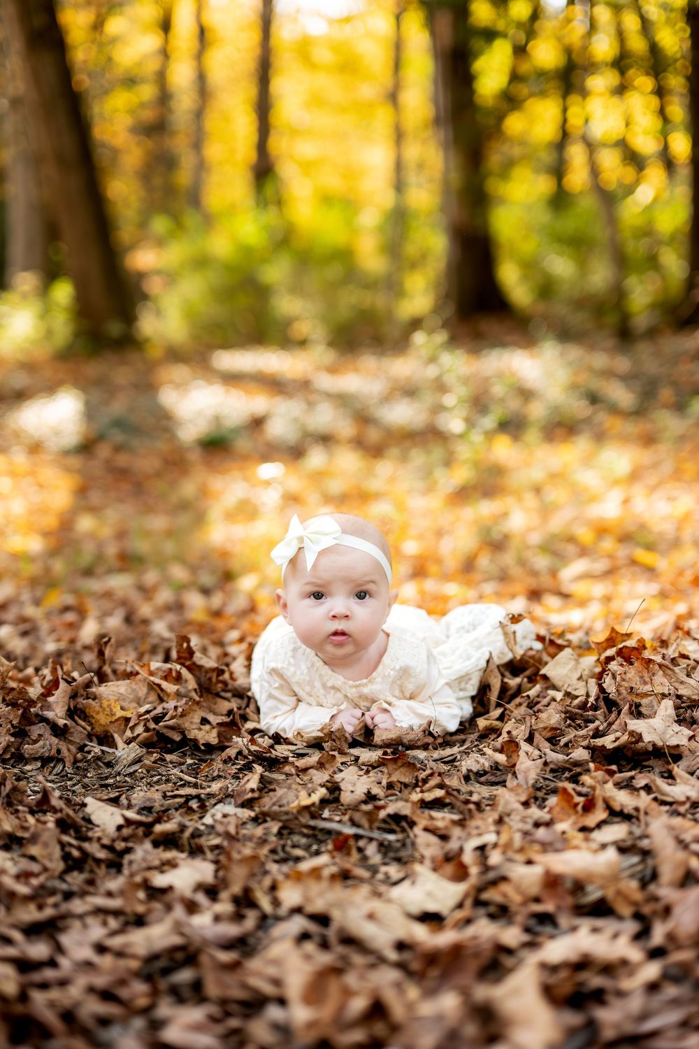 Baby girl in white lace outfit lying on a bed of fallen autumn leaves in a sunny forest.